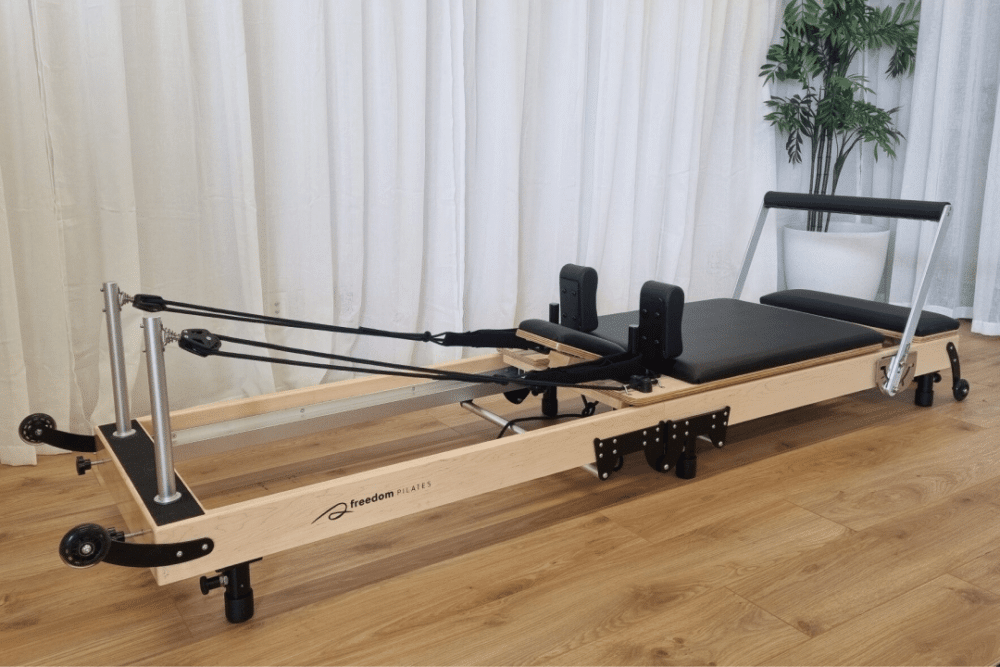 A black Soho, a foldable reformer with a light-coloured maplewood frame and black padding, sits on a wooden floor with a white curtain in the background.